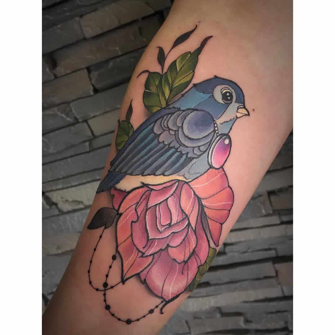 A colorful tattoo of a blue bird perched on a pink rose with green leaves and beads on an arm