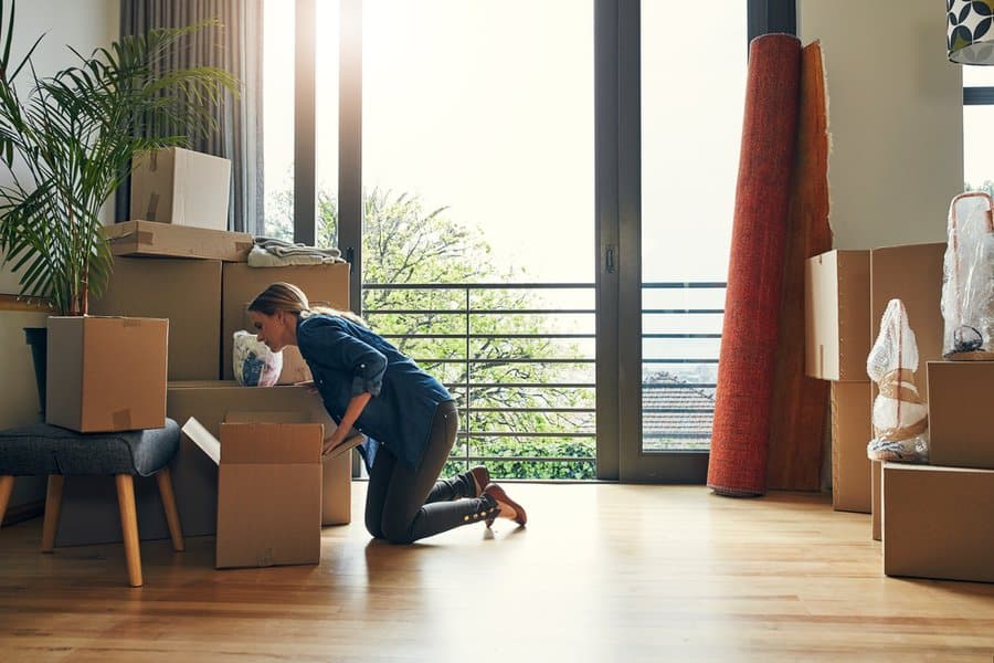 Woman kneeling on the floor, unpacking boxes in a bright room with large windows and moving supplies scattered around