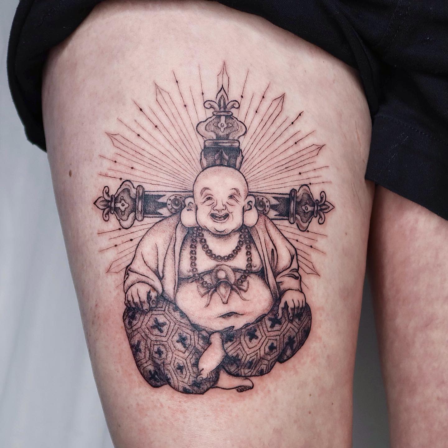 Black and white tattoo of a seated Buddha with a crown and rays on a thigh