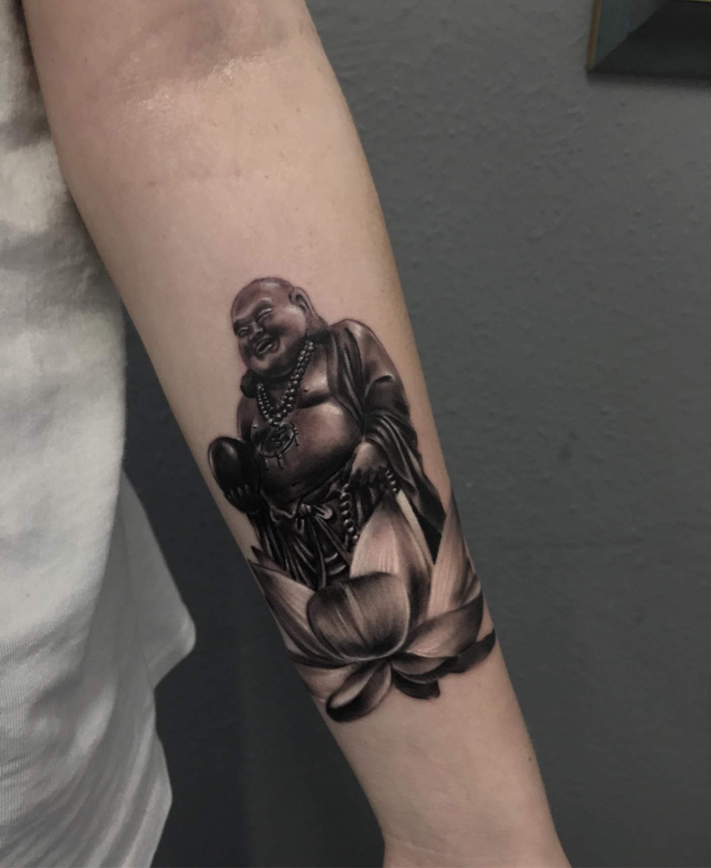 Black and gray tattoo of a laughing Buddha holding beads, surrounded by a lotus flower on forearm