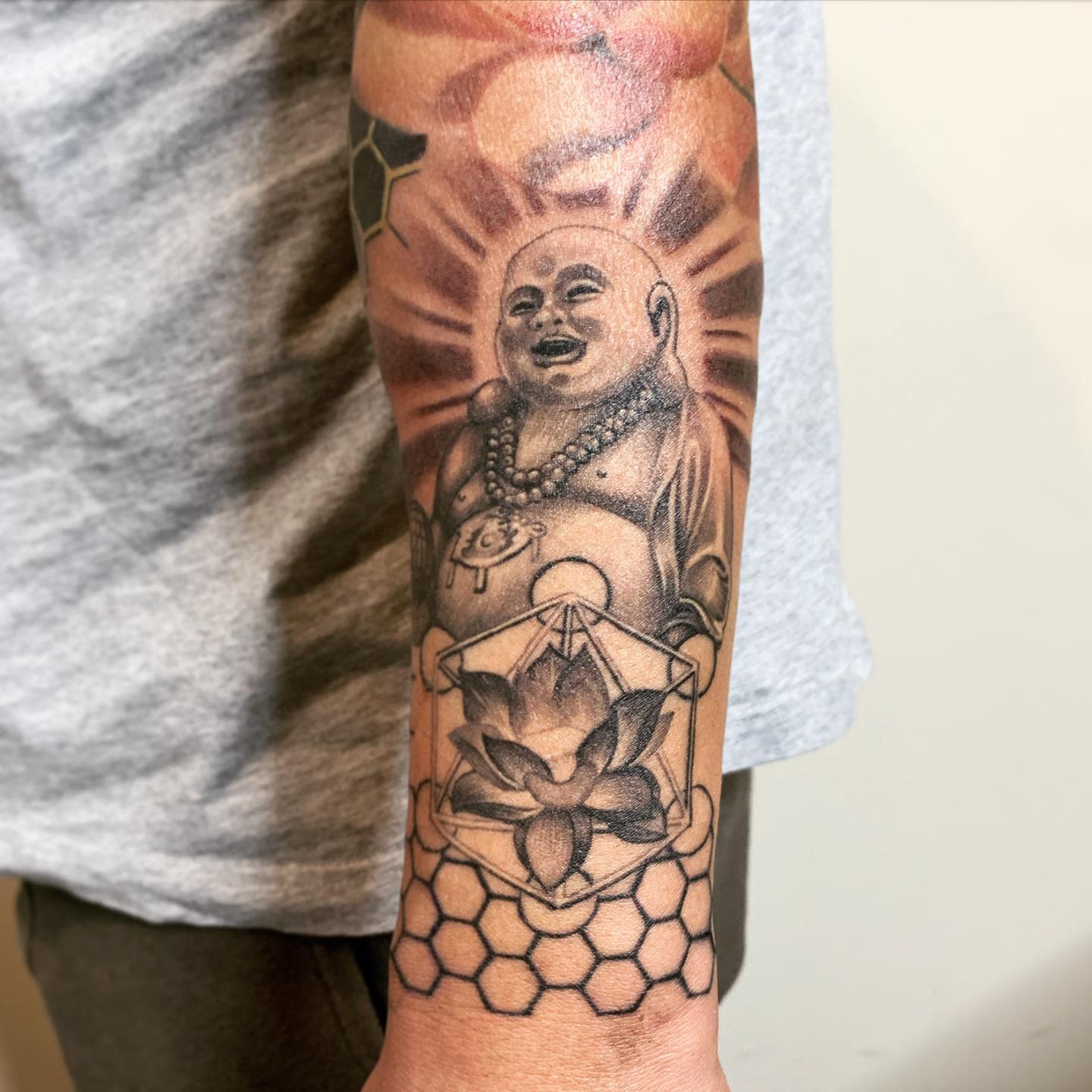 Forearm tattoo featuring a smiling Buddha, lotus flower, hexagonal pattern, and geometric shapes