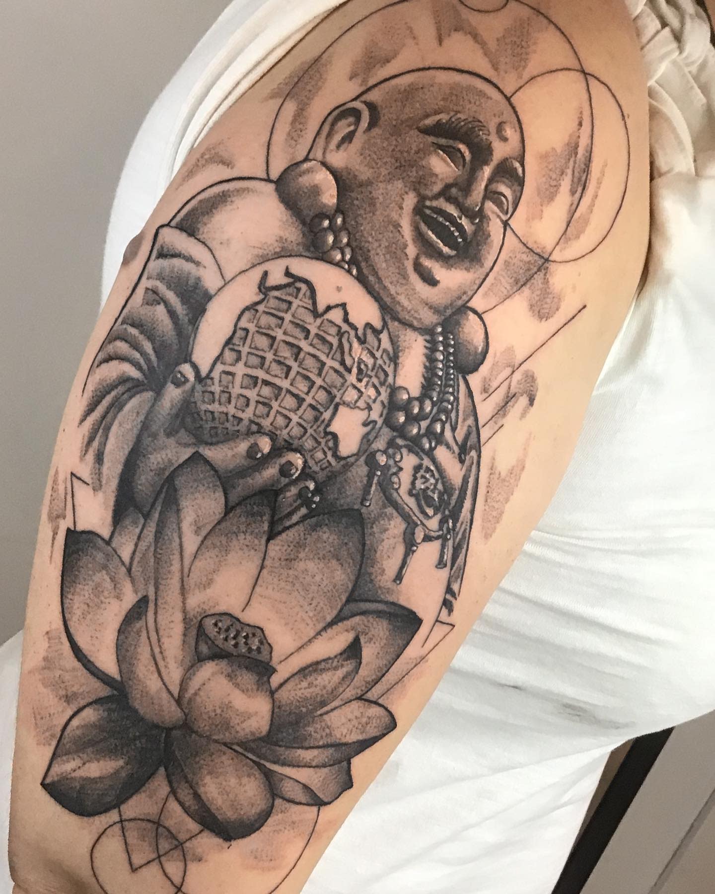 Tattoo of a laughing Buddha holding a globe, seated on a lotus flower, on an upper arm