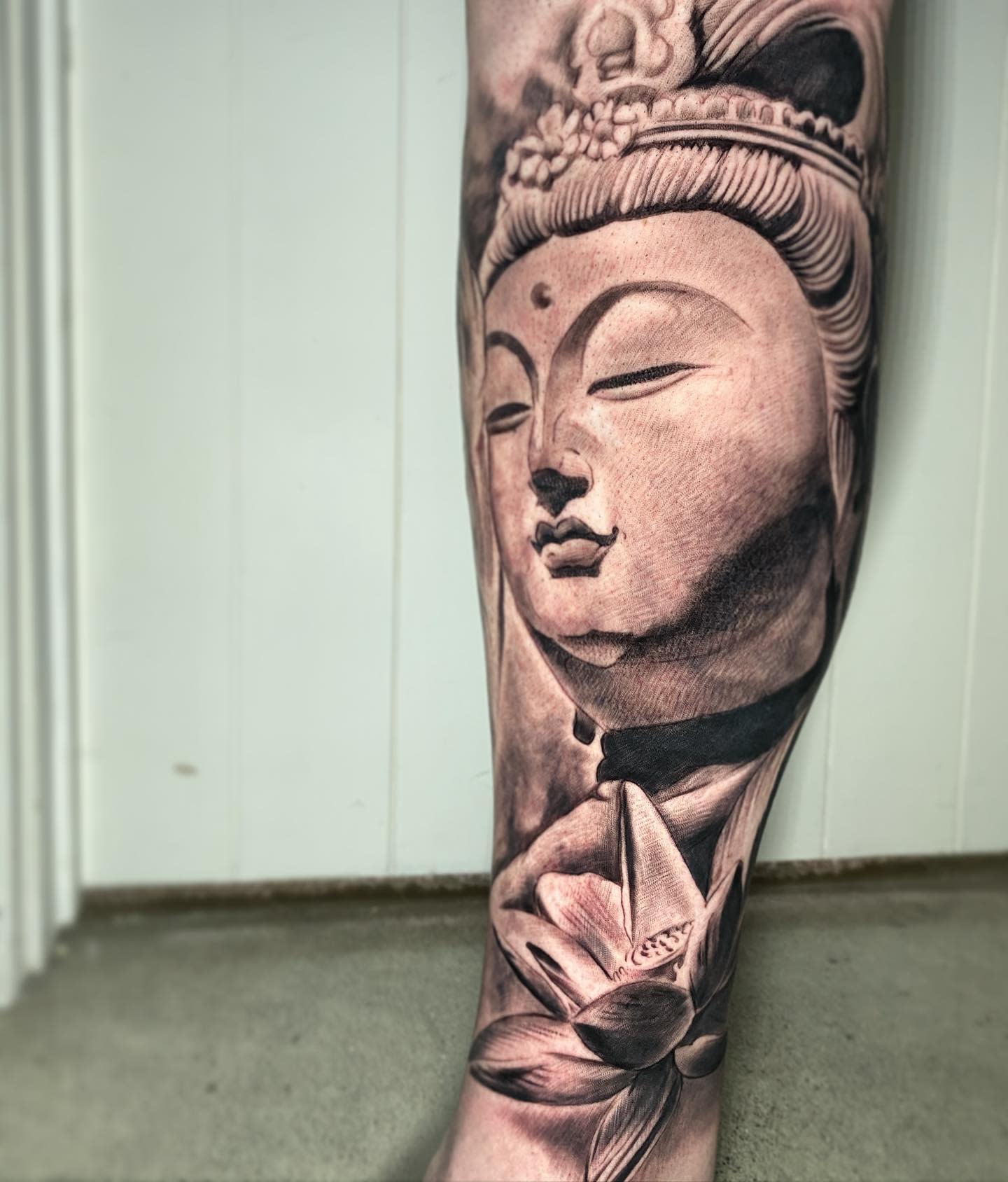 A black and gray tattoo of a serene Buddha face with a lotus flower on a leg