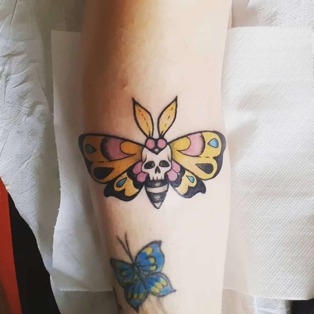 Colorful moth tattoo on the forearm, featuring bold yellow and pink wings with a skull at the center for a striking and gothic look.