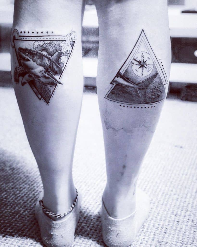 Ocean-themed leg tattoos featuring a shark breaking through waves inside a triangle and a compass with a shark fin on the other leg