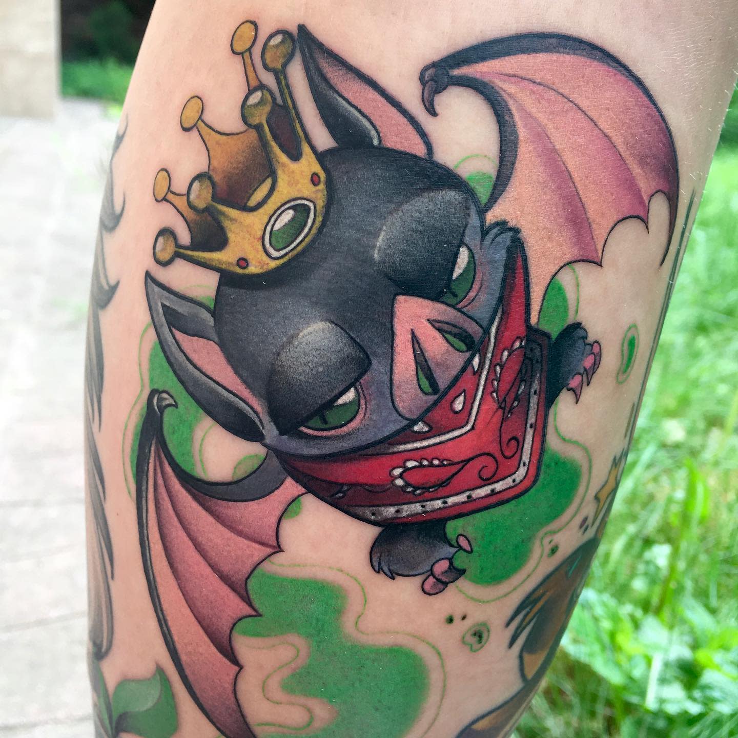 Tattoo of a bat wearing a crown and bandana, surrounded by green smoke on a forearm