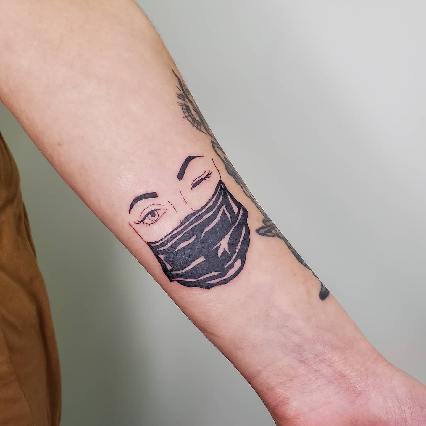 Tattoo of a winking face wearing a face mask on a forearm