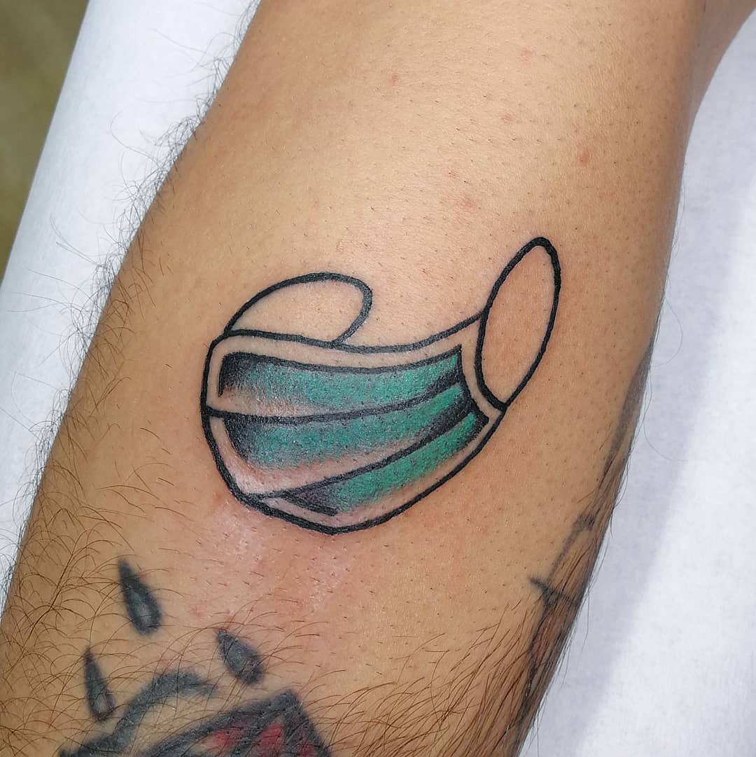Tattoo of a green and black face mask on an arm above the elbow