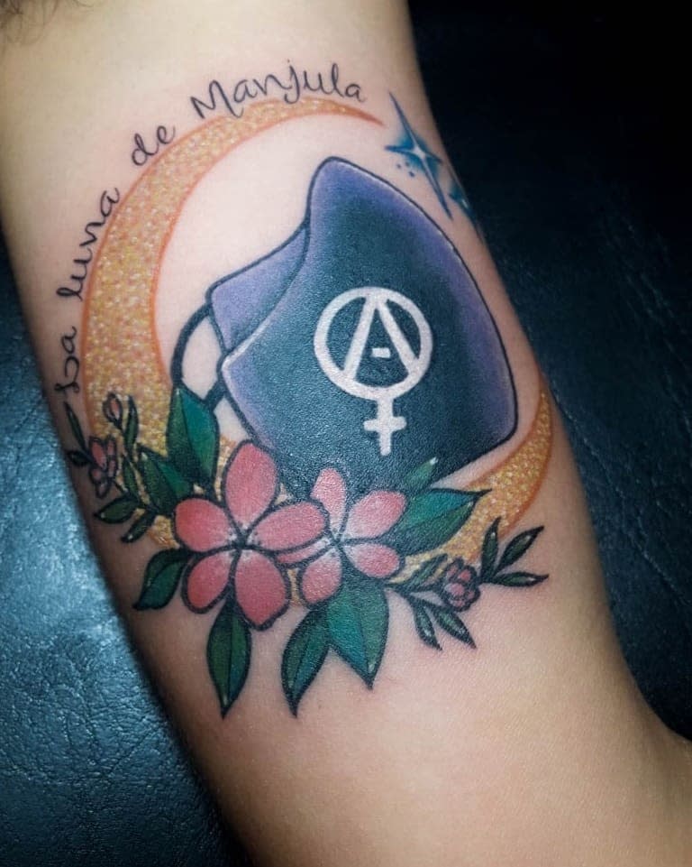 Tattoo of a facemask, pink flowers, yellow moon, and text "la luna de Maryula"