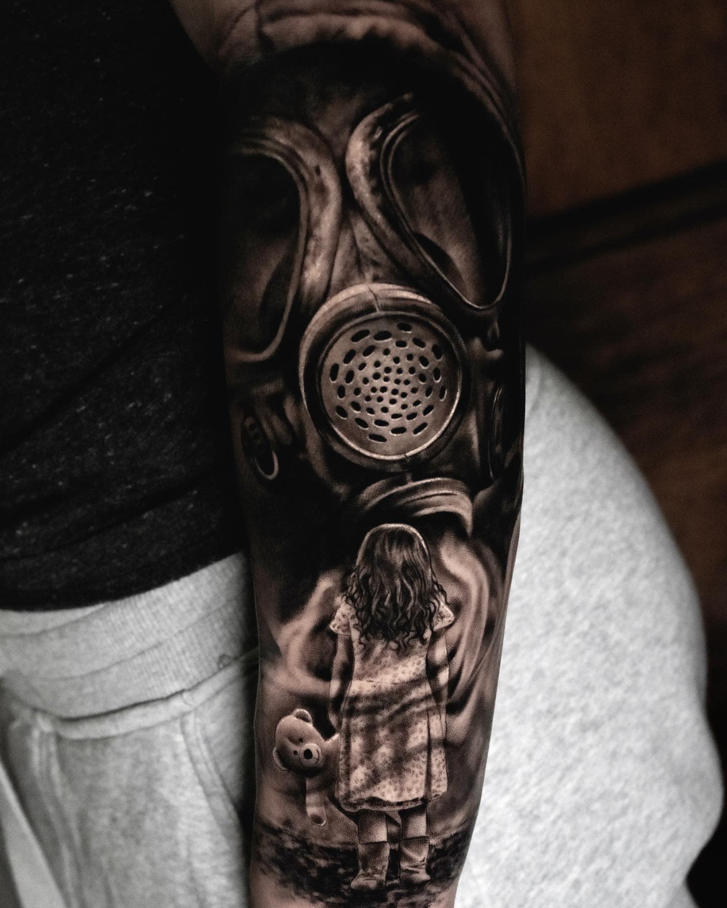 Tattoo of a girl holding a teddy bear facing a large gas mask on someone's arm