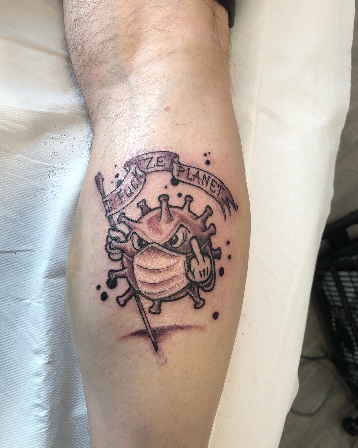 Tattoo of a masked virus cartoon holding up middle finger, with text "FUCK ZE PLANET
