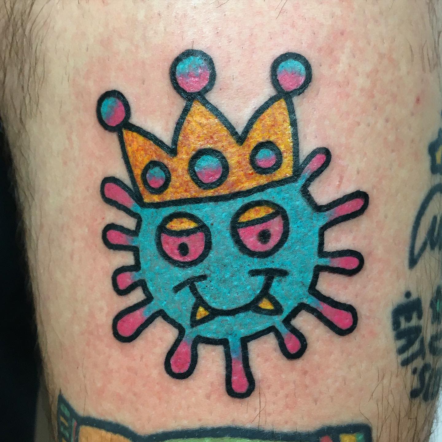 Tattoo of a blue virus cartoon with a yellow crown featuring pink and blue details