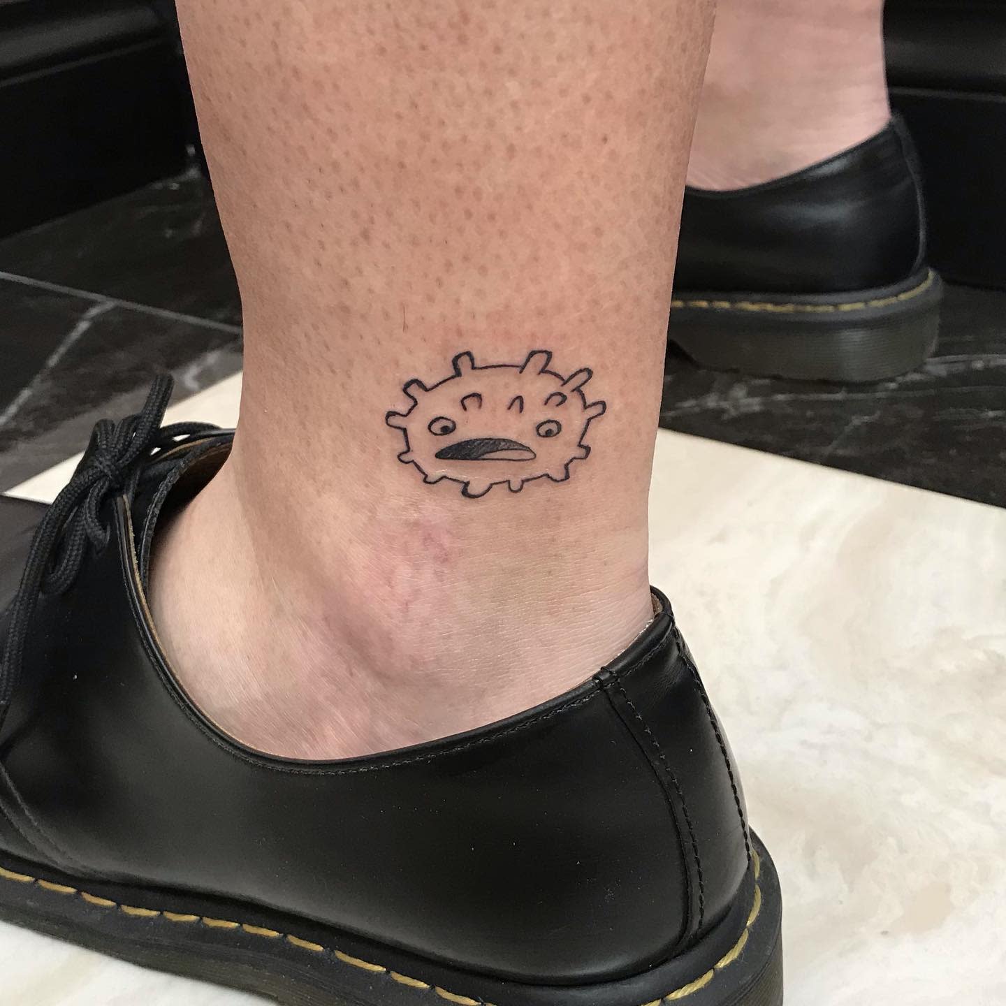 Tattoo of a sad virus-like character on the back of an ankle