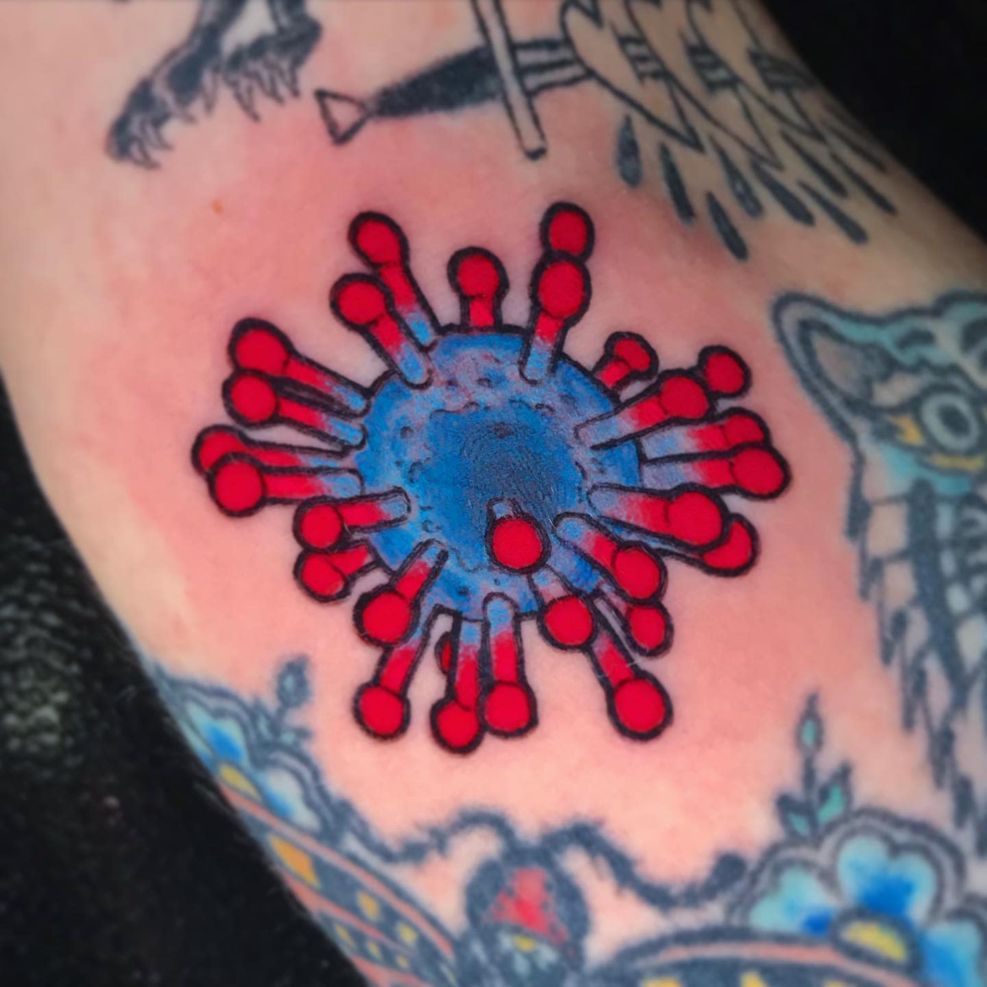 Tattoo of a blue and red virus symbol on an arm surrounded by other tattoos
