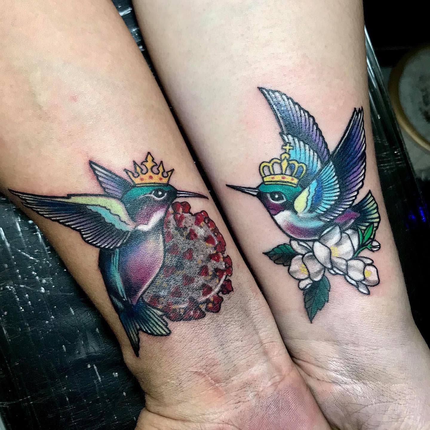 Two arms with matching hummingbird tattoos; one with a crown and virus, the other with flowers