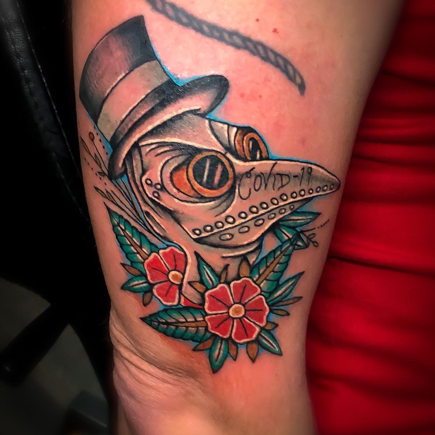 A tattoo of a plague doctor with "COVID-19" text, flowers, and leaves above knee