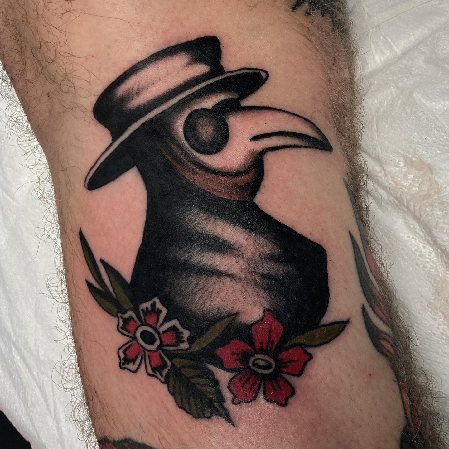 Tattoo of a plague doctor surrounded by red and white flowers on skin