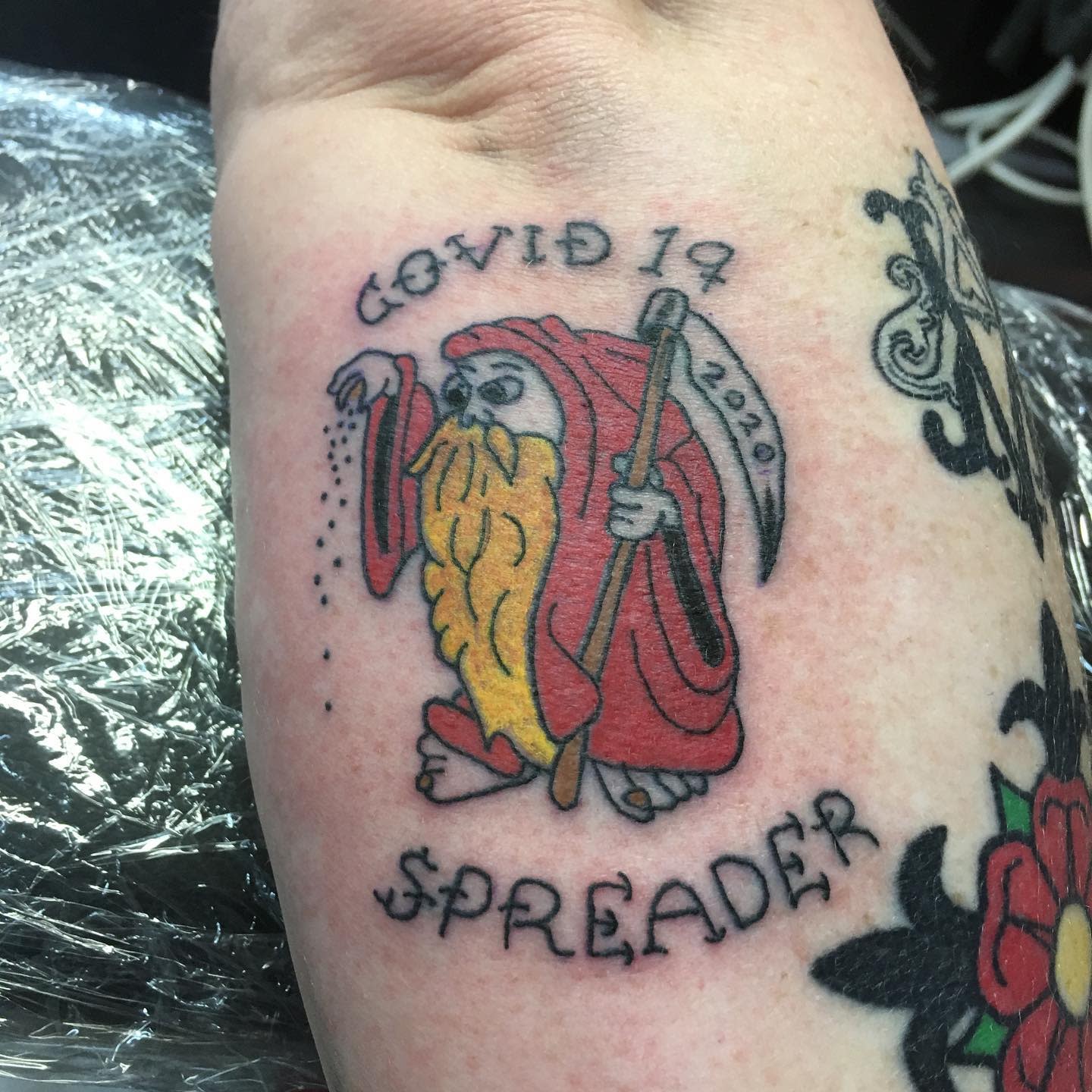 Tattoo of a red-cloaked Grim Reaper labeled "Covid 19 Spreader" on an upper arm