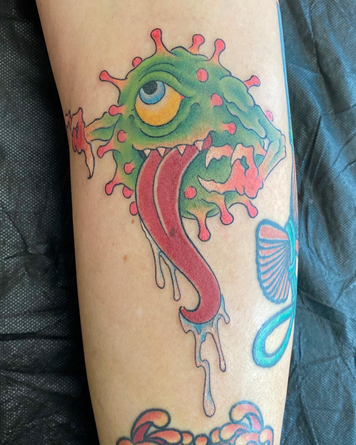 Tattoo of a one-eyed green monster with red tongue and pink splatters on skin