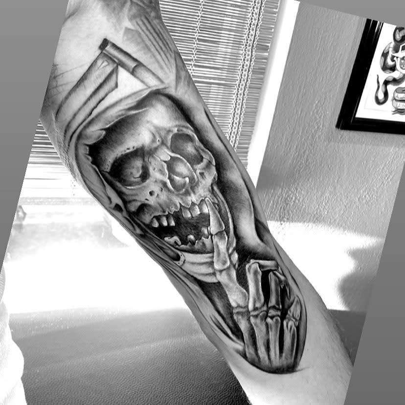 Black and white tattoo of a skull with a finger over its mouth on an arm
