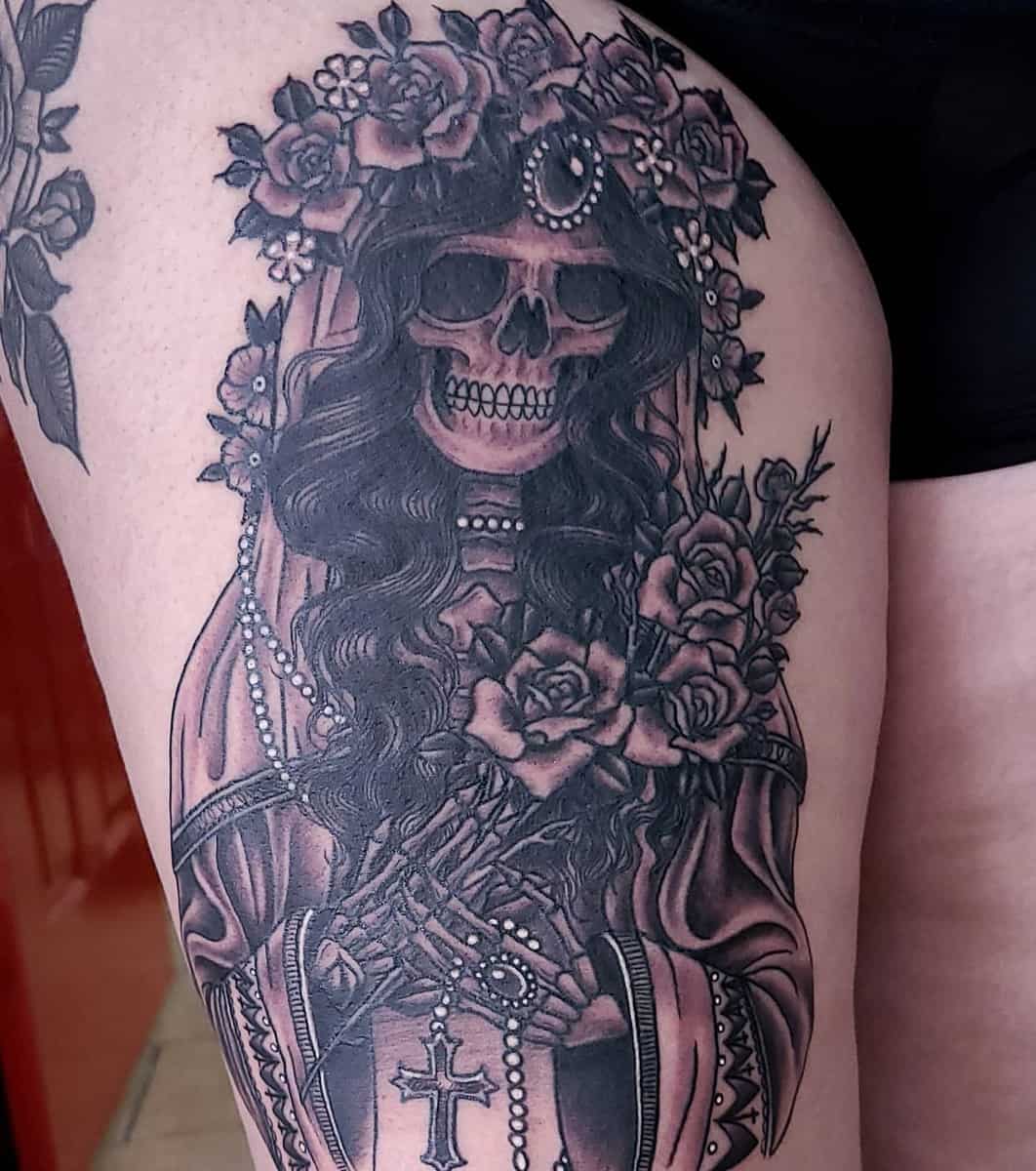 Santa Muerte thigh tattoo with a floral crown, holding a cross, and adorned in pearls and roses