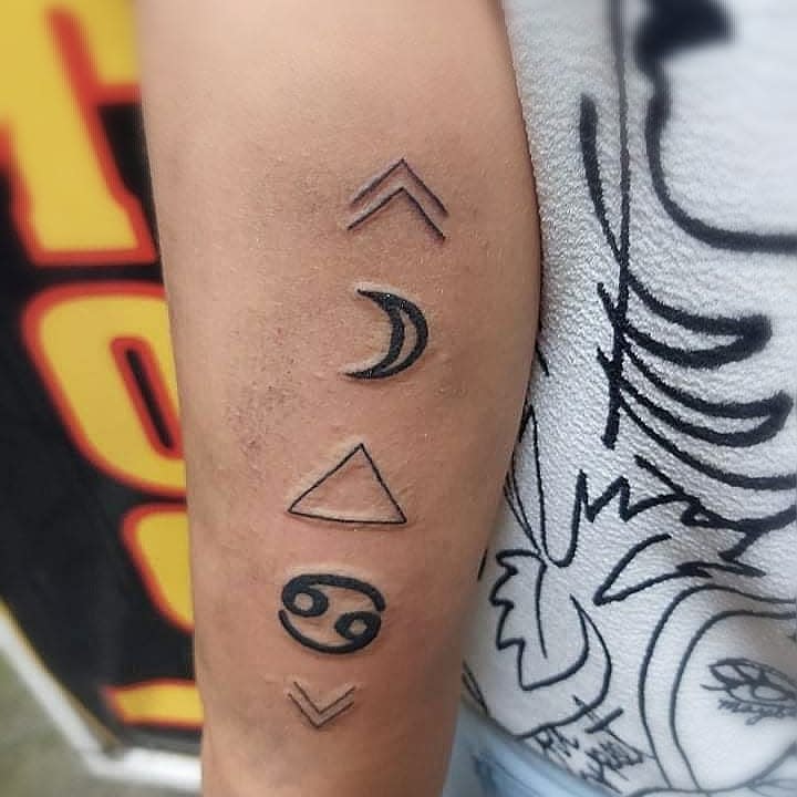 Tattoo of various symbols on an arm including arrows, a moon, a triangle, and a Cancer zodiac sign