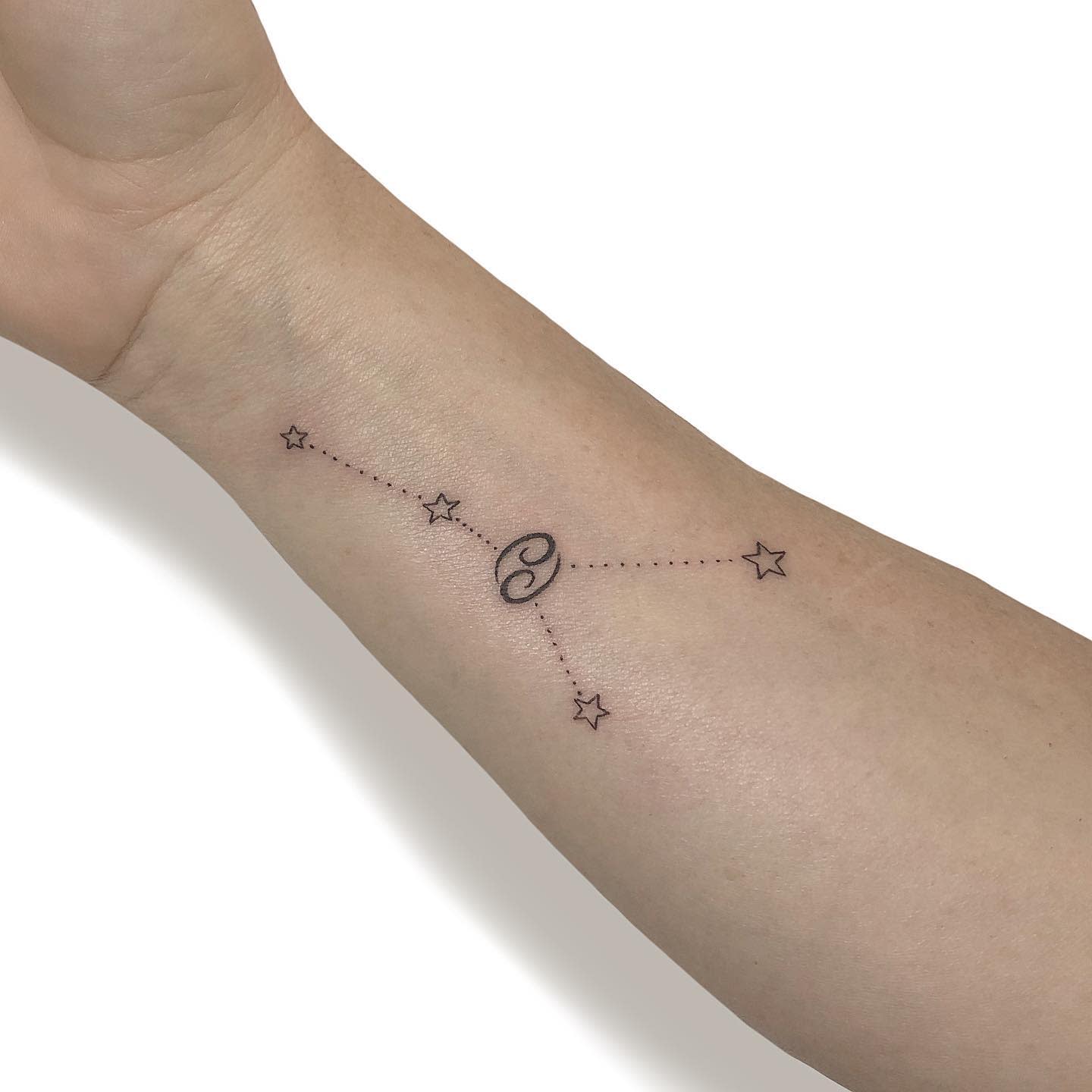A forearm tattoo featuring the Cancer zodiac symbol connected to stars by dotted lines