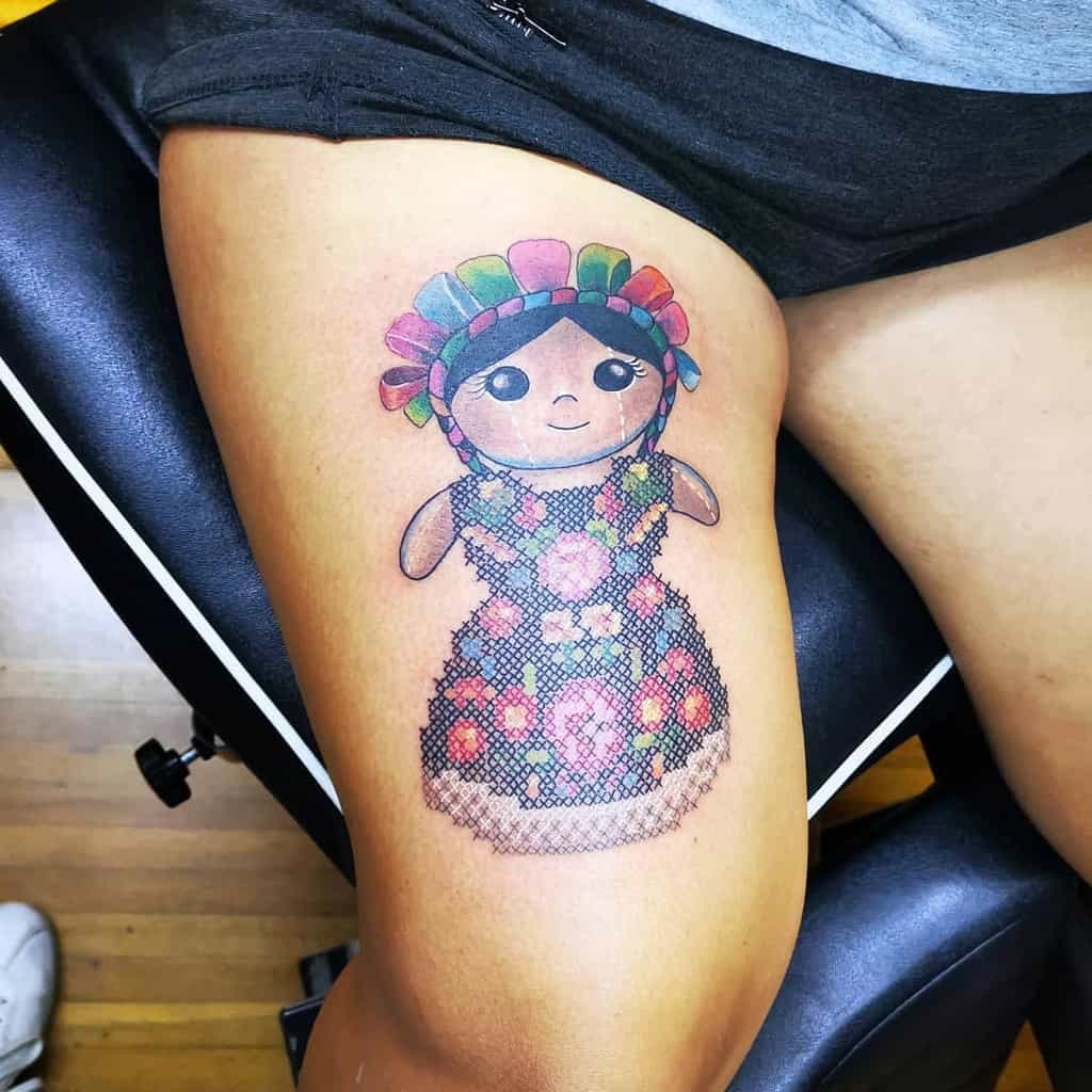 Vibrant cross-stitch style tattoo of a traditional Mexican doll with a colorful headdress, blending folk art with embroidery-inspired design