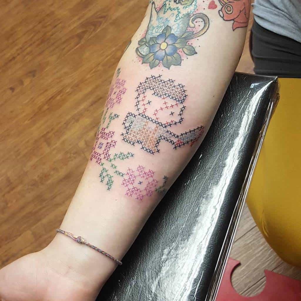 Whimsical cross-stitch style tattoo of a human holding a stick, surrounded by floral patterns