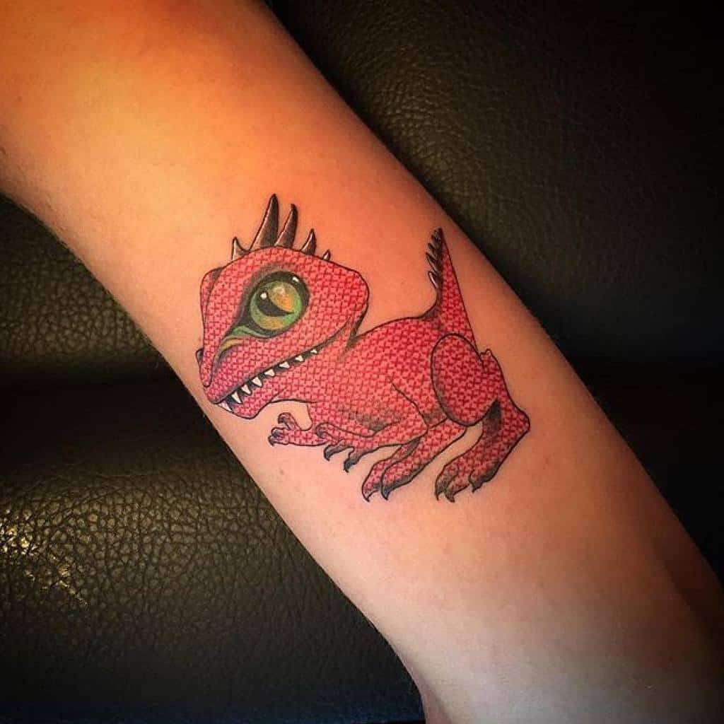 Playful cross-stitch style tattoo of a red baby dinosaur with a large green eye, blending embroidery texture with bold cartoon details
