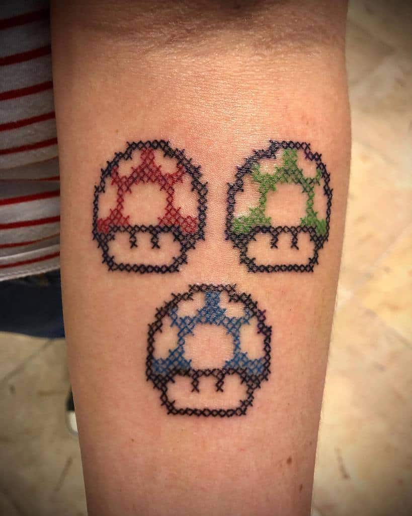 Cross-stitch style tattoo of three pixelated Super Mario mushrooms in red, green, and blue