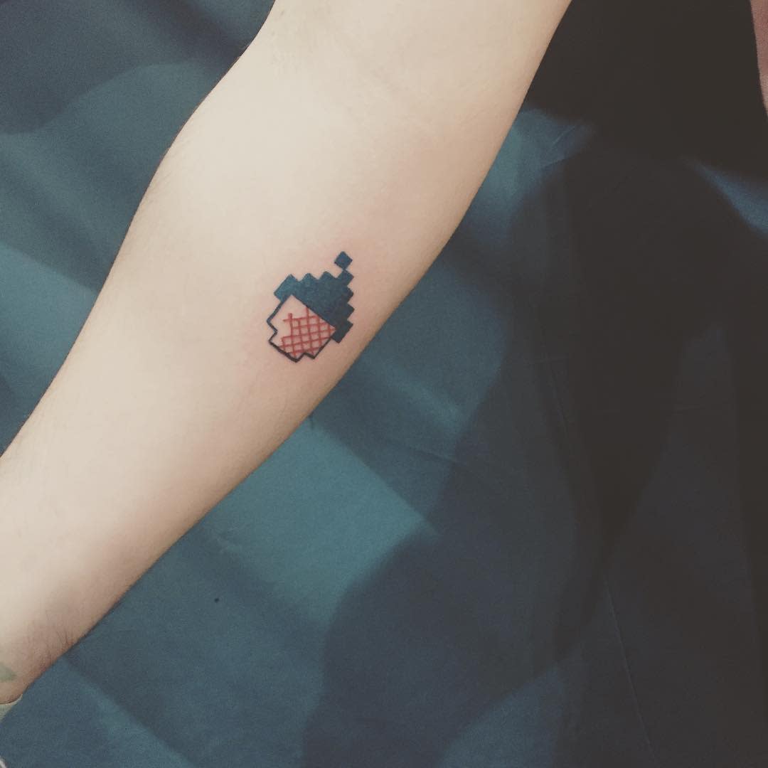 Minimalist cross-stitch style tattoo of a pixelated acorn in red and blue, blending digital aesthetics with embroidery-inspired design
