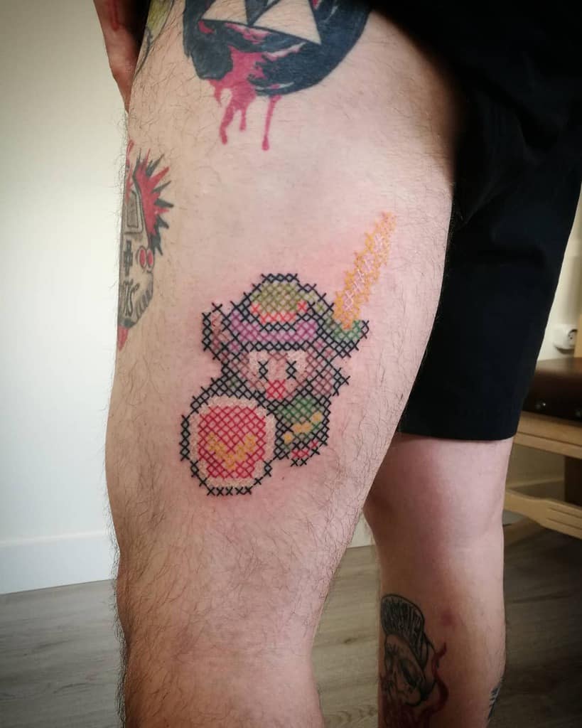 Cross-stitch style tattoo of Link from The Legend of Zelda, featuring pixelated embroidery details with a sword and shield design