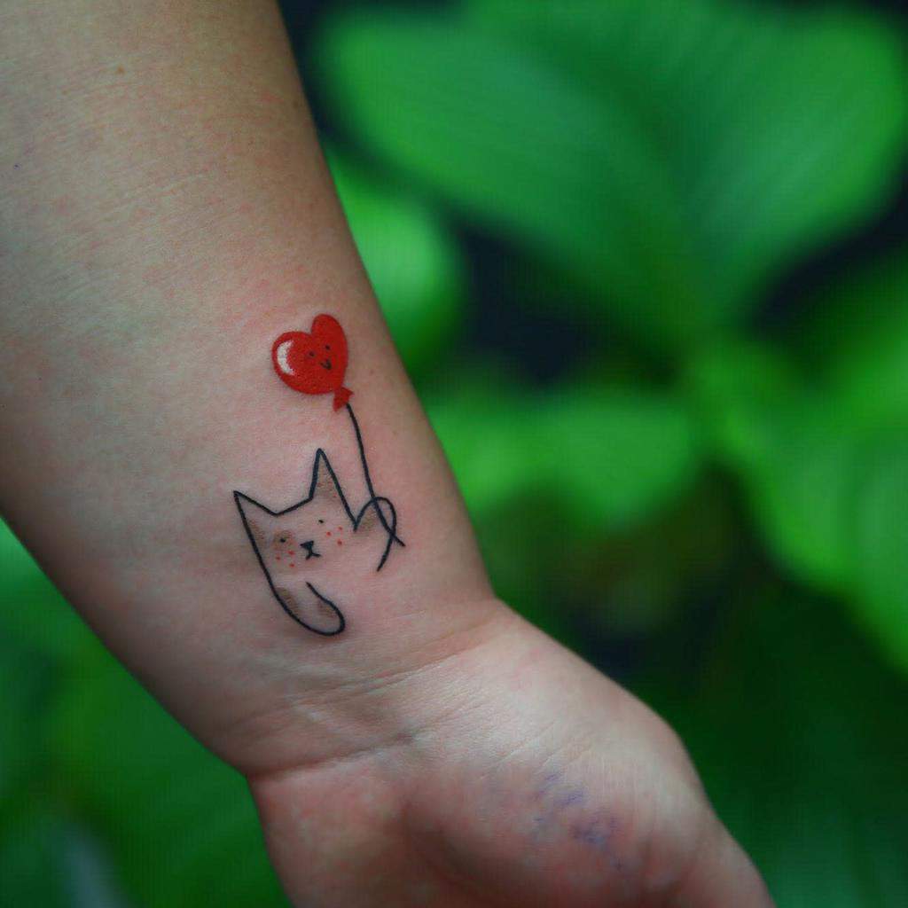 Minimalist cat tattoo clutching a red heart balloon on a wrist