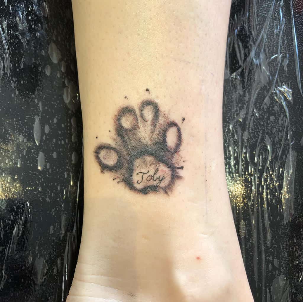 Tattoo of a cat paw print with the name "Toby" in the center on the back of a person's ankle