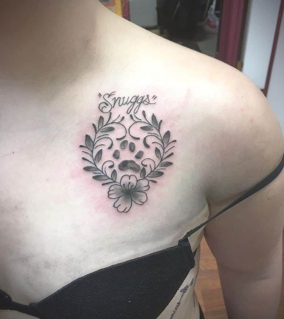 A tattoo with a cat paw print, leaves, and a flower on someone's chest, with the word "Snuggs" above it