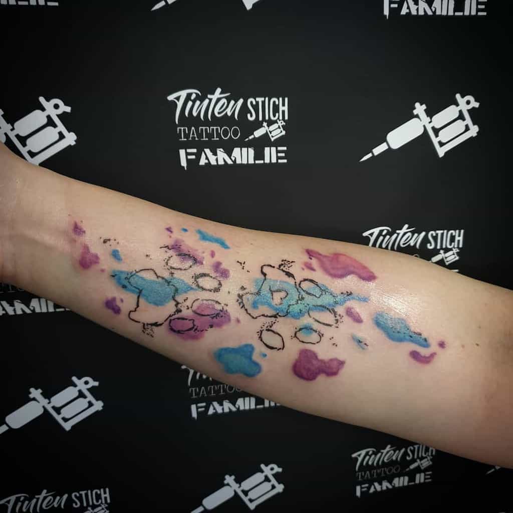 Forearm tattoo with an abstract watercolor design in blue, purple, and black ink, featuring a subtle cat paw for a touch of playful charm