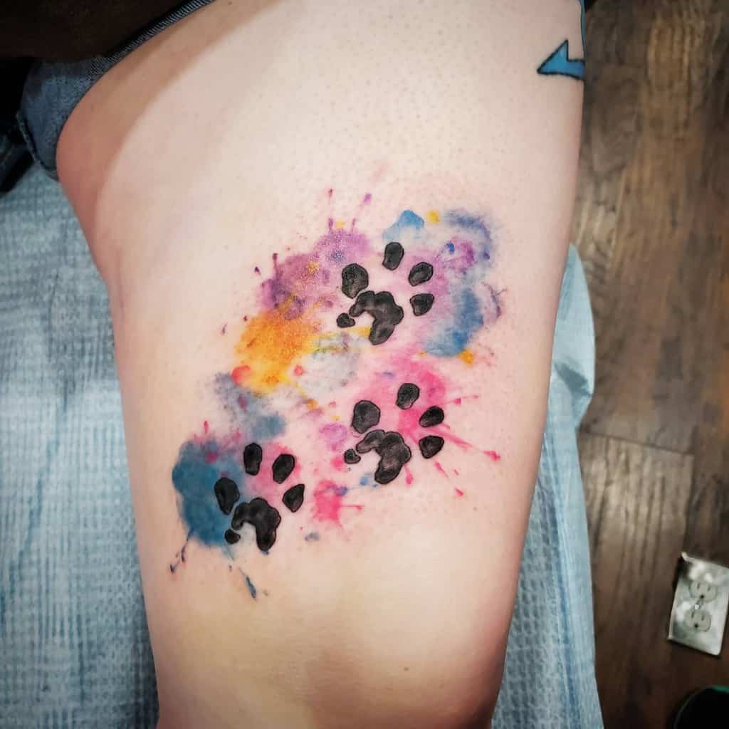 Colorful cat paw print tattoos on a person's thigh, with splashes of purple, pink, yellow, and blue