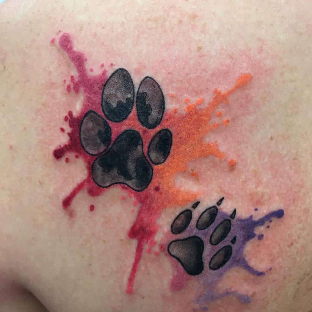 Two black cat paw print tattoos with colorful paint splatters on a person's skin