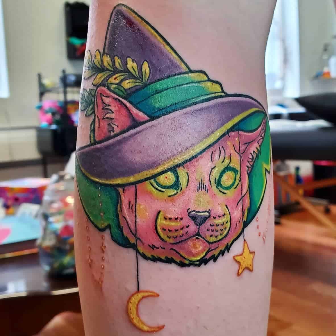 Tattoo of a cat in a witch hat, with a crescent moon and star hanging from the brim