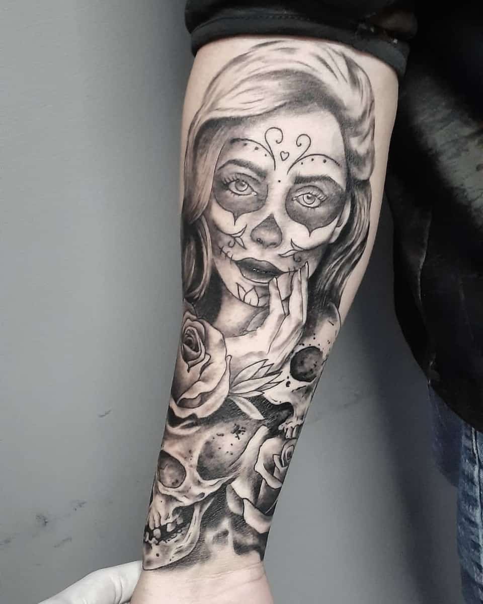 Realistic tattoo of a woman with Catrina makeup, holding her hand to her lips, adorned with roses and skulls