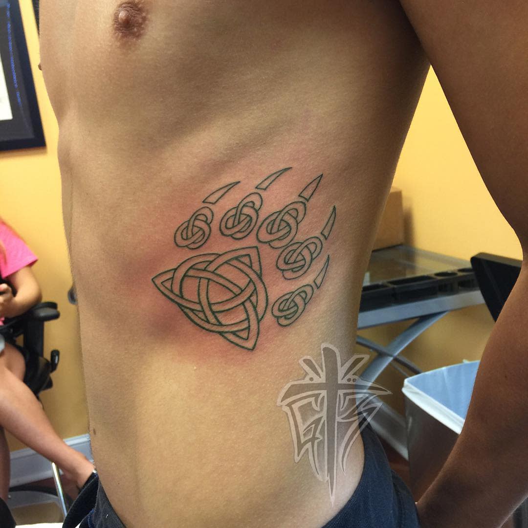Tattoo of a bear paw with a Celtic knot on a person's side torso, in a room with yellow walls