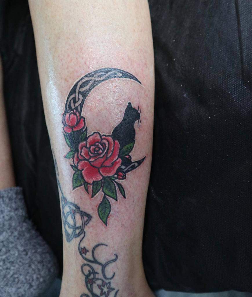 Tattoo of a black cat perched on a crescent moon, surrounded by vibrant red roses and lush green leaves on a person's arm