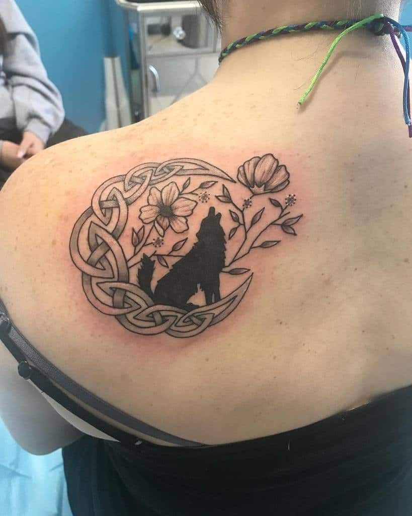 Tattoo of a howling wolf with flowers, a crescent moon, and Celtic knot design on a person's upper back