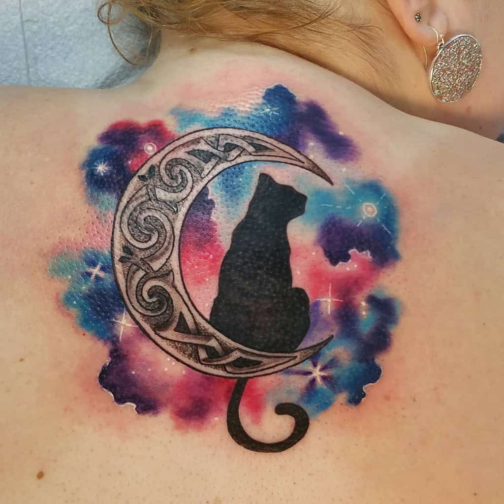 Tattoo of a cat perched on a crescent moon with colorful galaxy hues, elegantly inked on a person’s upper back