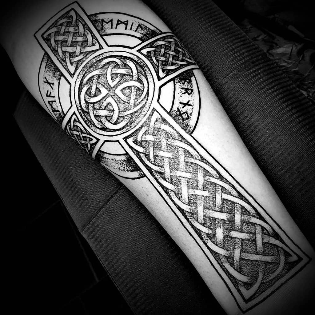 A detailed black and white Celtic cross tattoo on the forearm, featuring intricate knotwork and a textured circular design with runic inscriptions