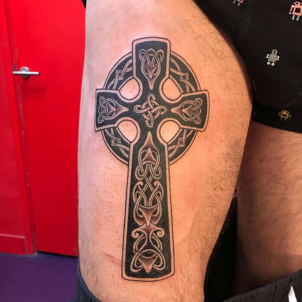 A detailed Celtic cross tattoo on the thigh, featuring intricate knotwork patterns and bold outlines, creating a striking and symbolic design