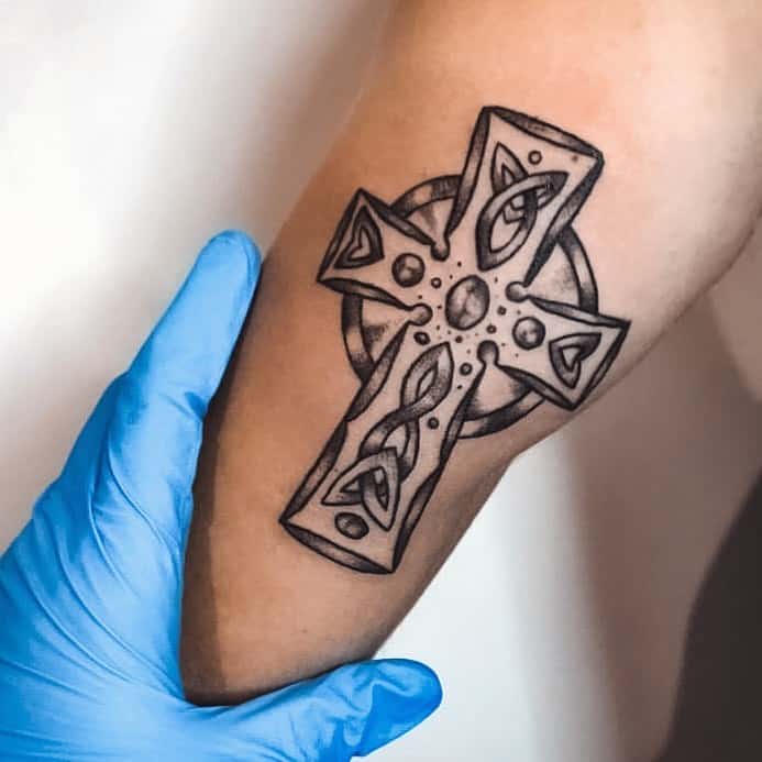 A Celtic cross tattoo on the upper arm, featuring bold lines, knotwork details, and dot accents, creating a modern yet traditional design