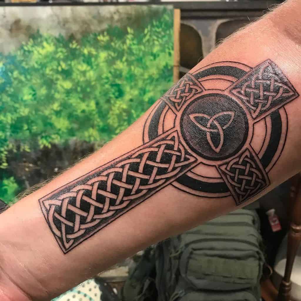 A detailed Celtic cross tattoo on the forearm, featuring intricate knotwork and a triquetra symbol in the center, surrounded by bold circular lines