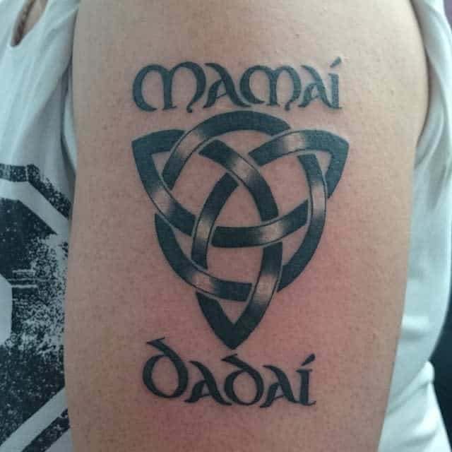 Celtic knot tattoo on arm with "Mamái" above and "Dadaí" below, in black ink, on light skin